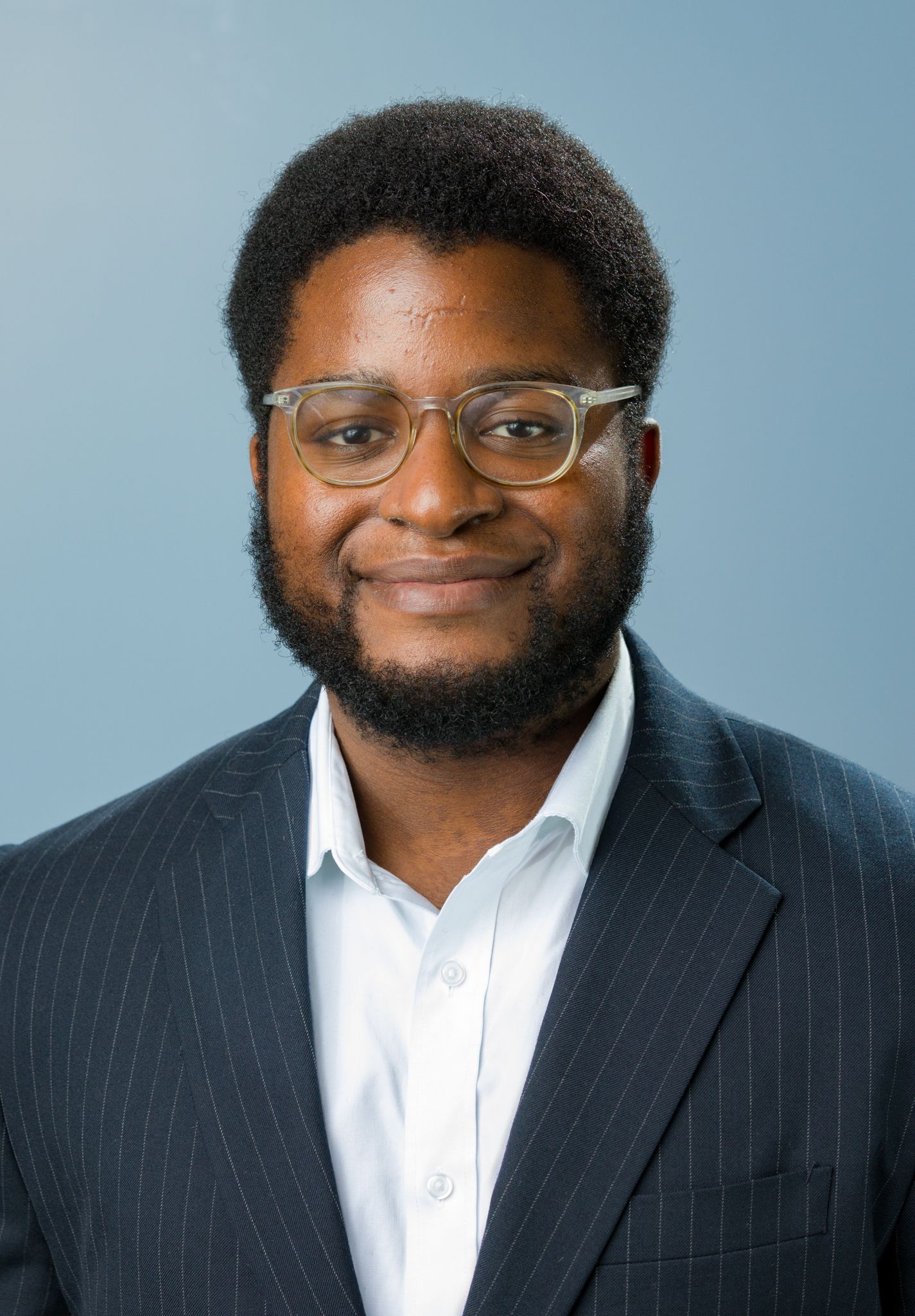 Professional headshot of a bearded man wearing glasses and a navy pinstripe suit on blue background.