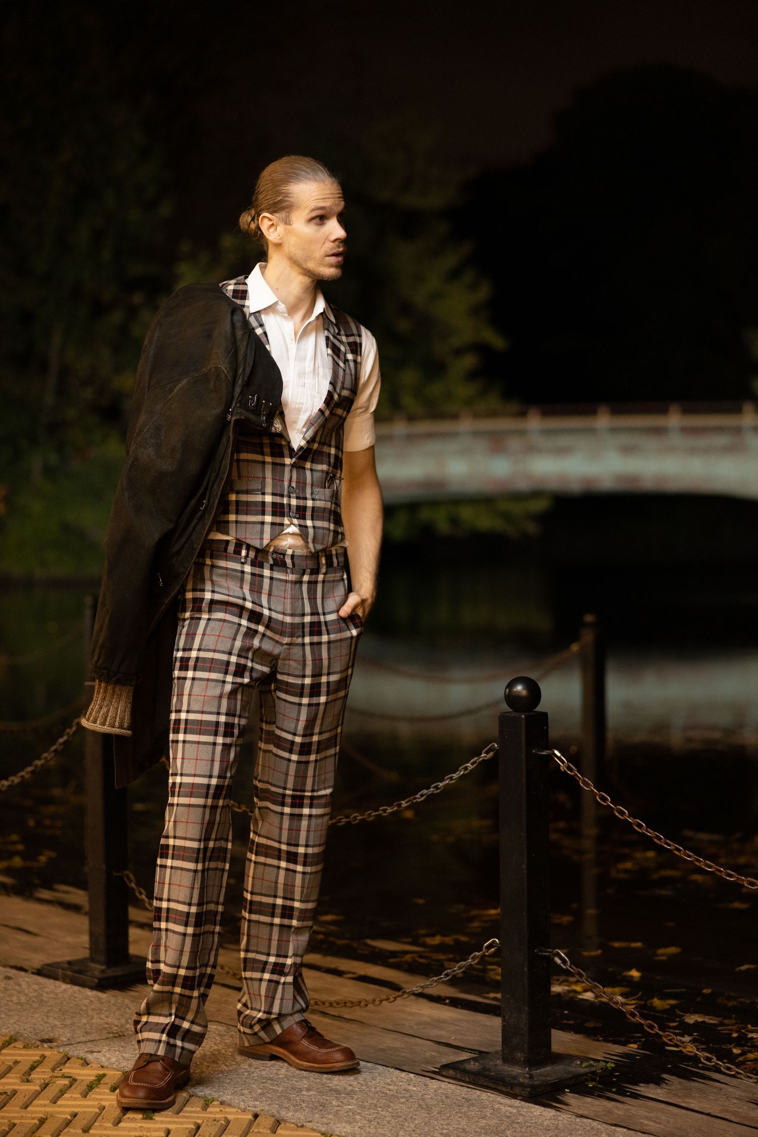 Tall man in plaid trousers and dark coat standing near chain posts by pond at night.