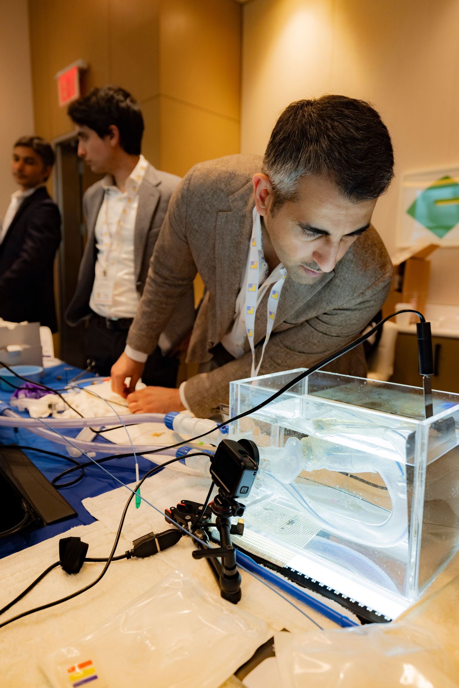 Man in blazer closely examining a illuminated aquarium device at an indoor conference exhibition table.