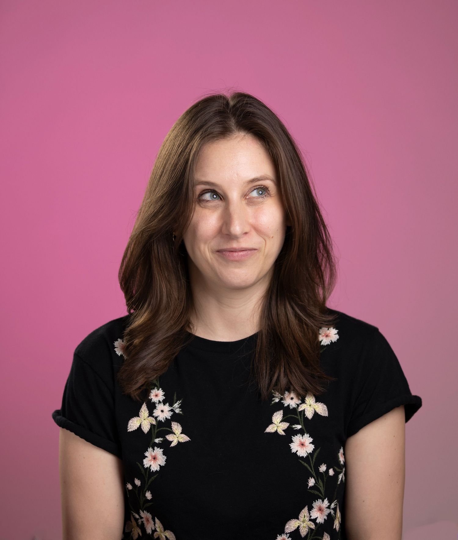 Brunette woman in a floral black top smiling against a bright pink background.
