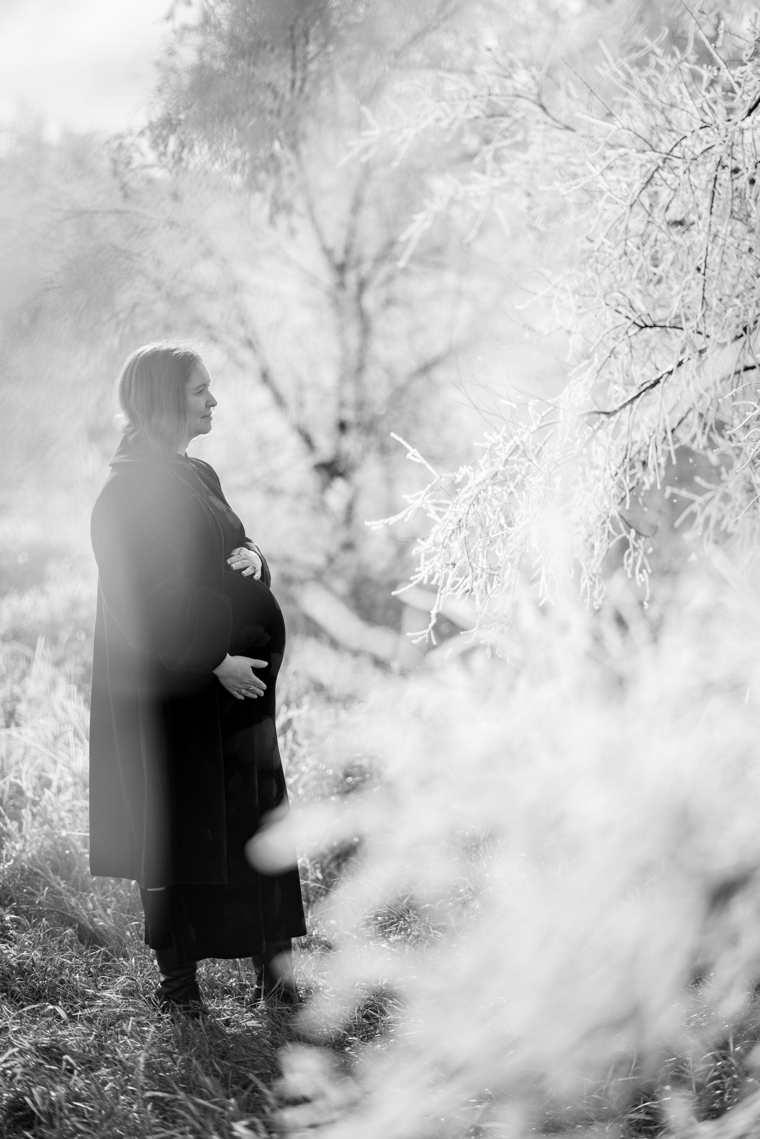 Black and white portrait of a pregnant woman in a long coat standing in a misty winter landscape.