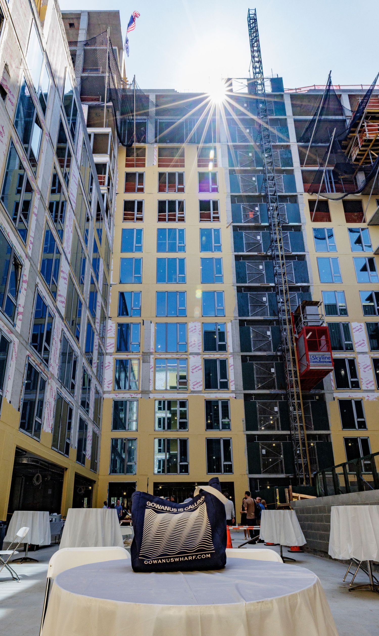 Construction site of a modern glass and concrete building with sun flare and crane visible against blue sky.