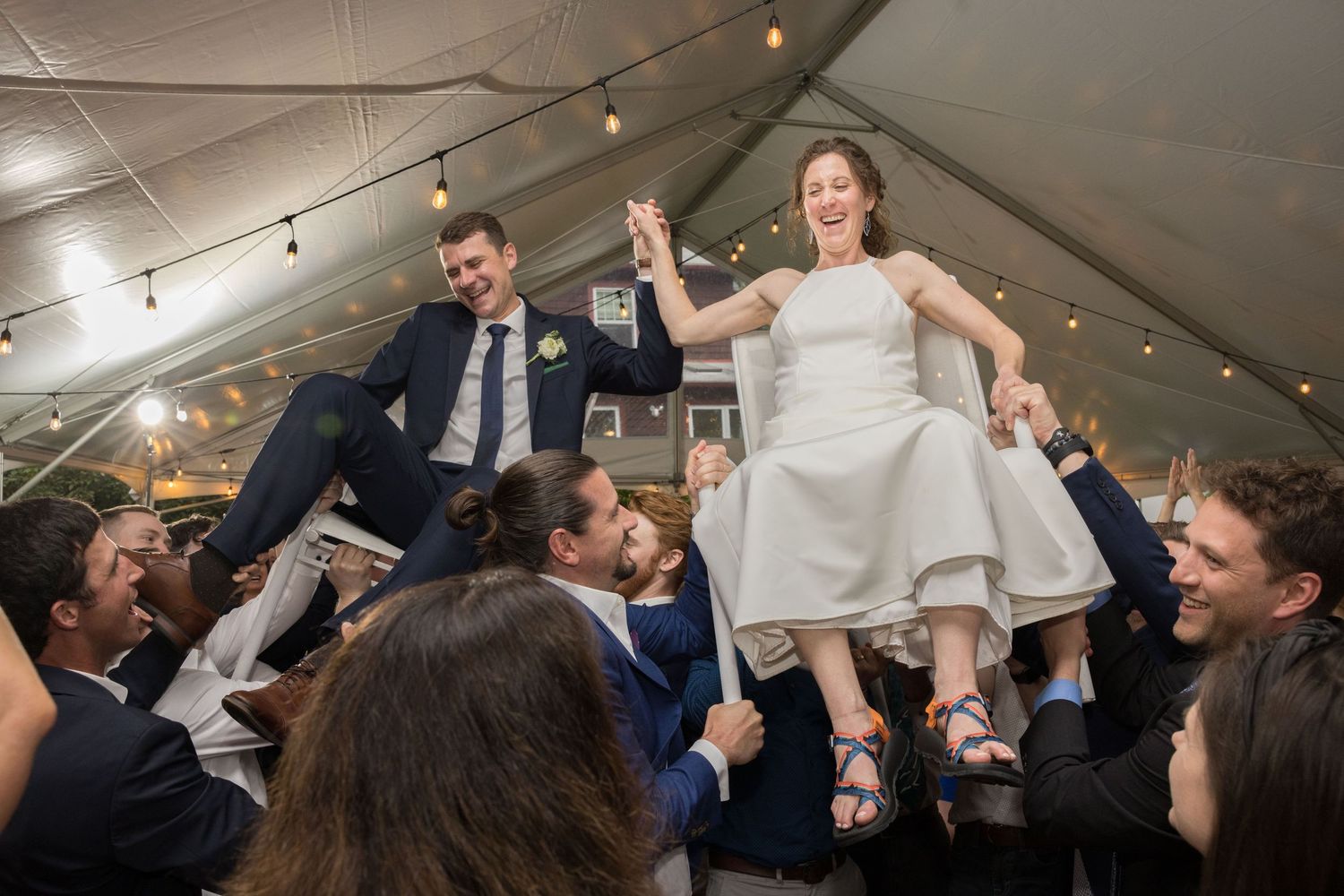 Bride and groom lifted by guests during joyful hora dance at elegant tented wedding reception celebration.