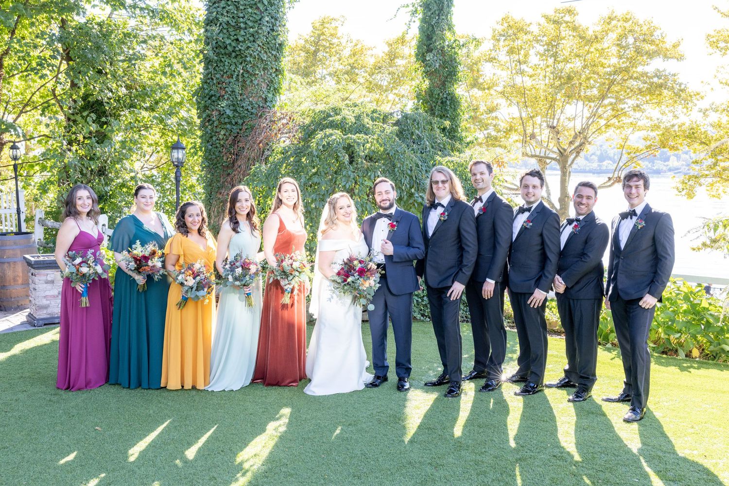 Wedding party poses in garden with bridesmaids in colorful gowns and groomsmen in black tuxedos.