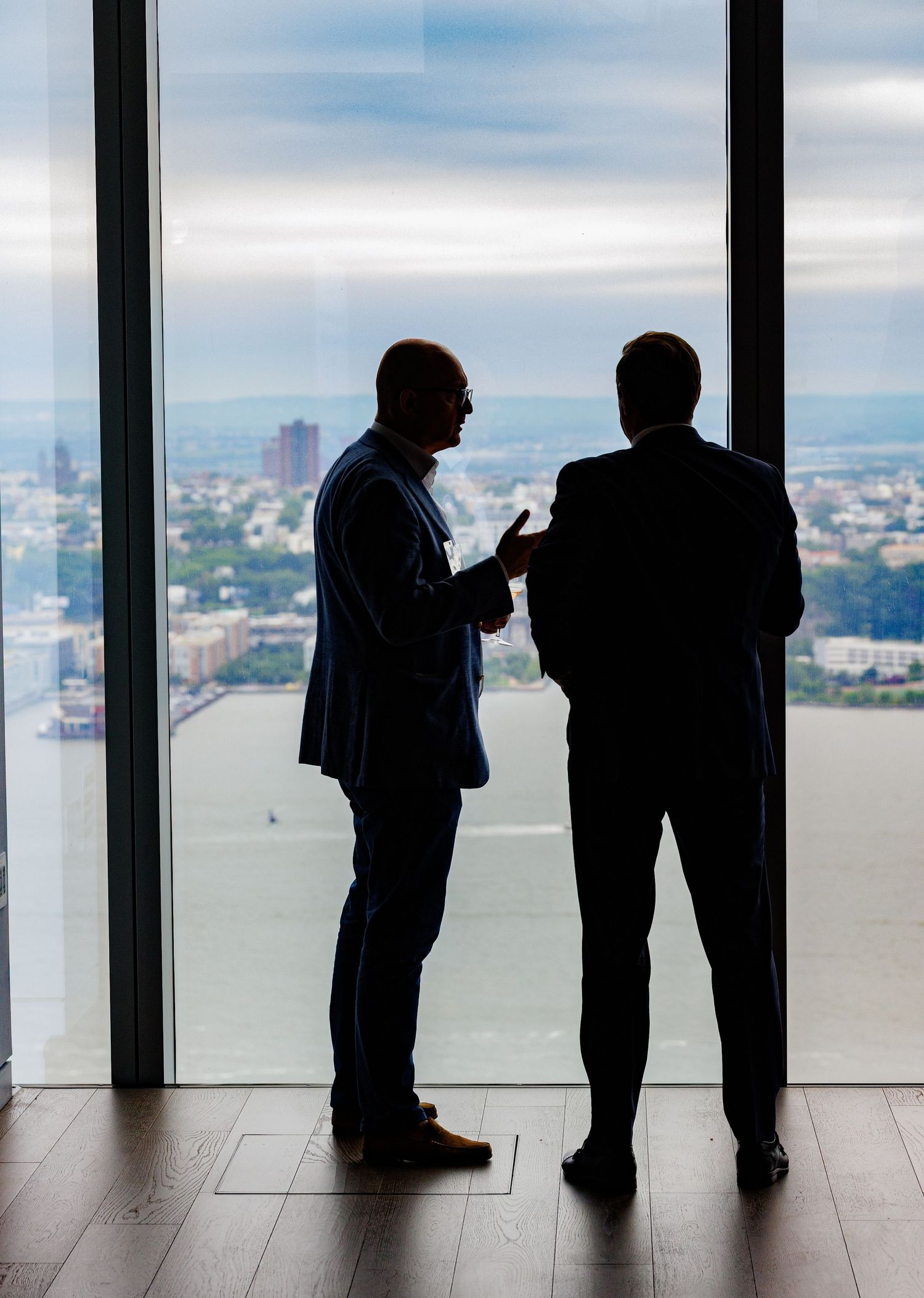 Two business professionals in silhouette conversing by floor-to-ceiling windows overlooking a city waterfront.