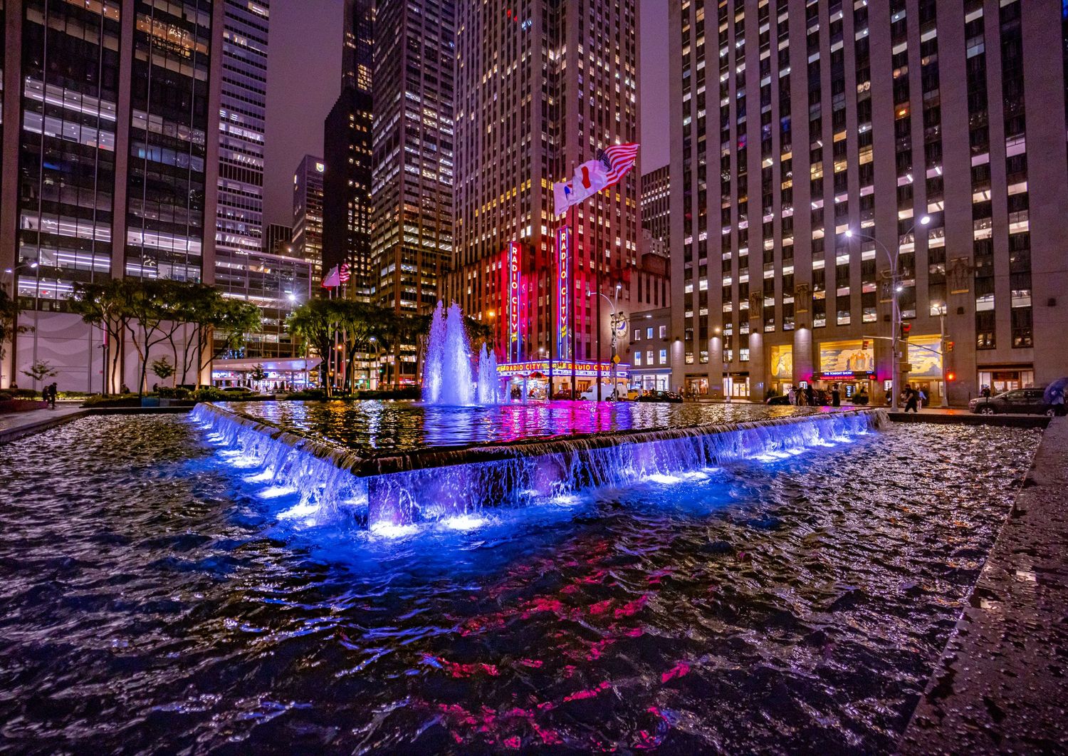 Illuminated blue fountains and waterfalls at Rockefeller Center plaza surrounded by skyscrapers at night in NYC.