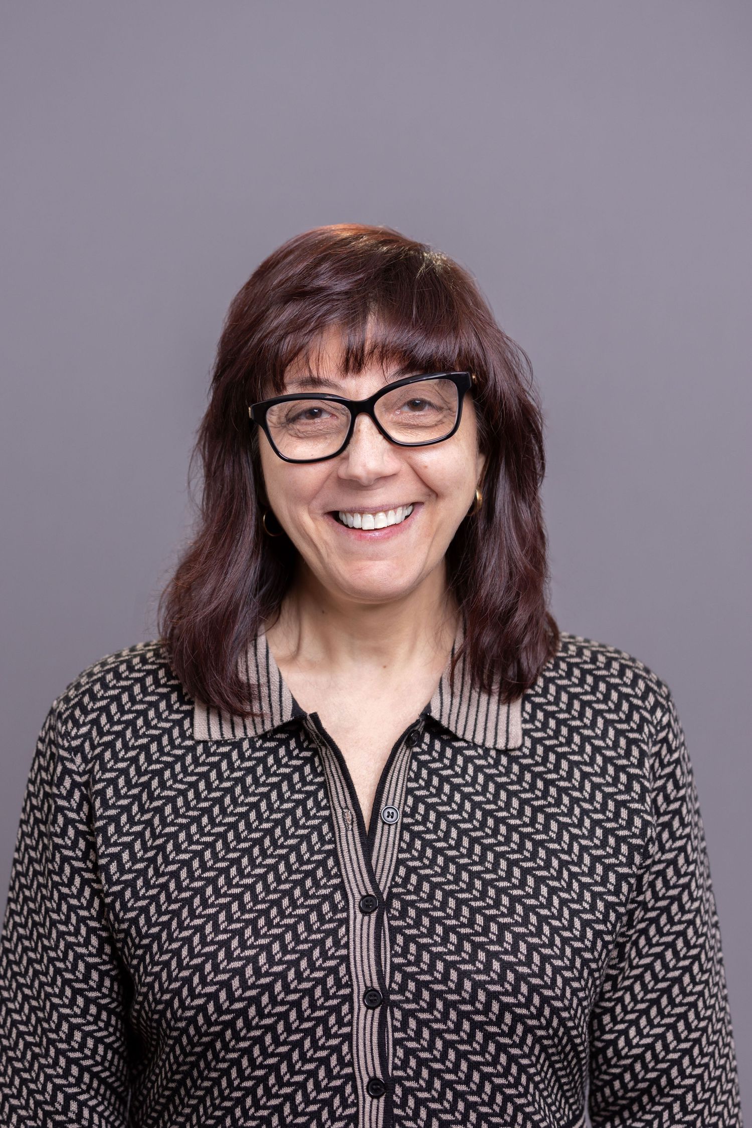 Professional headshot of a smiling woman with glasses and dark hair on gray background.