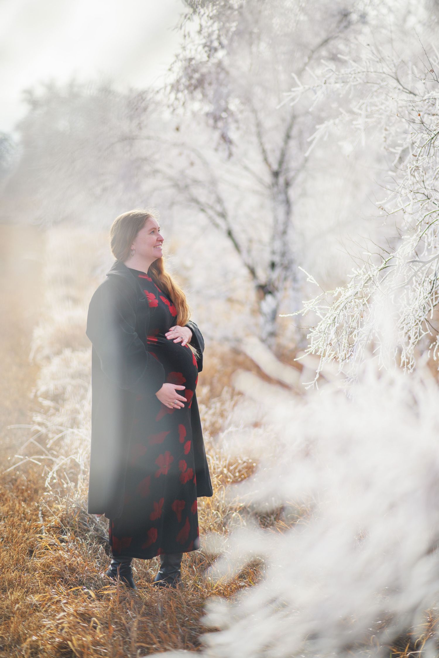 Pregnant woman in a red floral dress and dark coat standing in a sunlit frost-covered winter field.