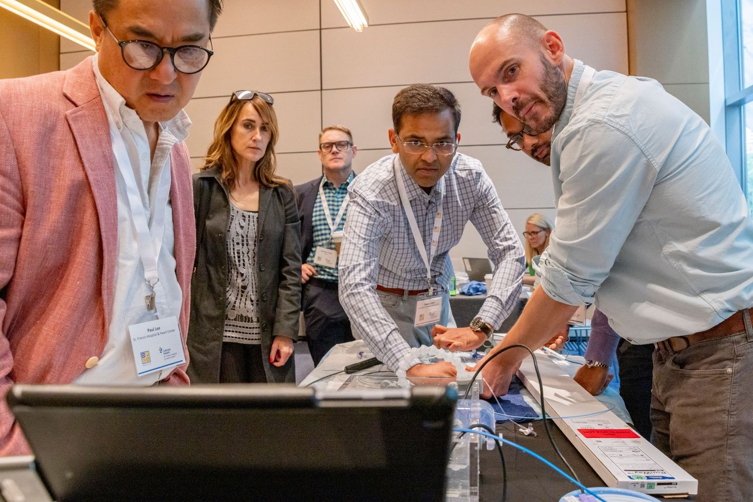 Group of conference attendees in badges gathered around a table examining technology demonstration indoors.