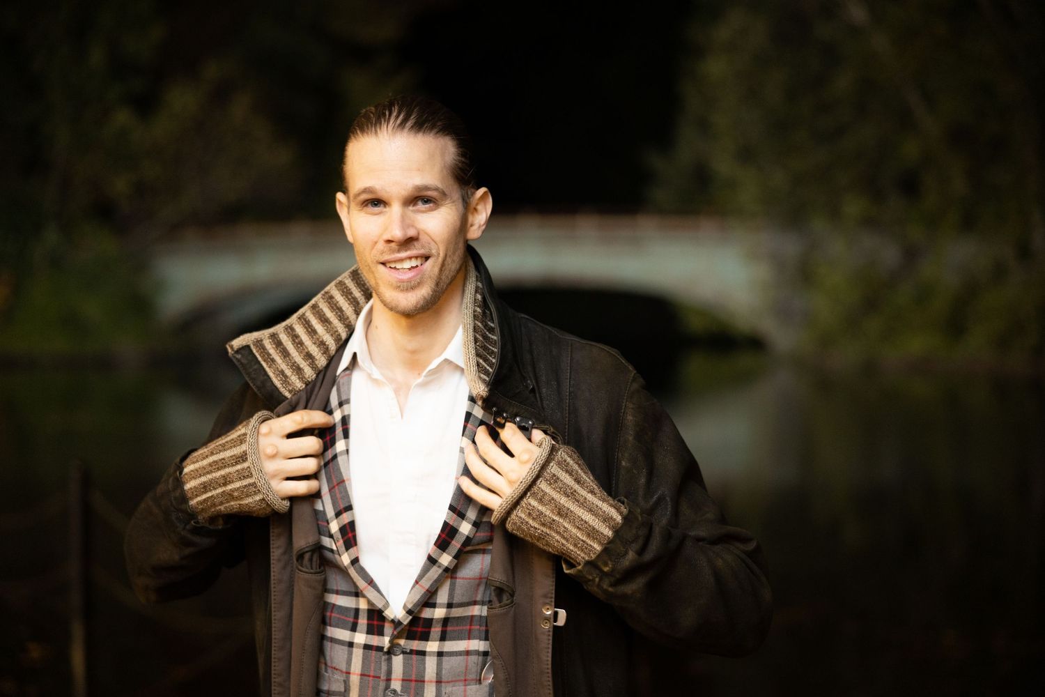 Smiling man in plaid blazer and leather jacket posing near bridge in dark outdoor setting.