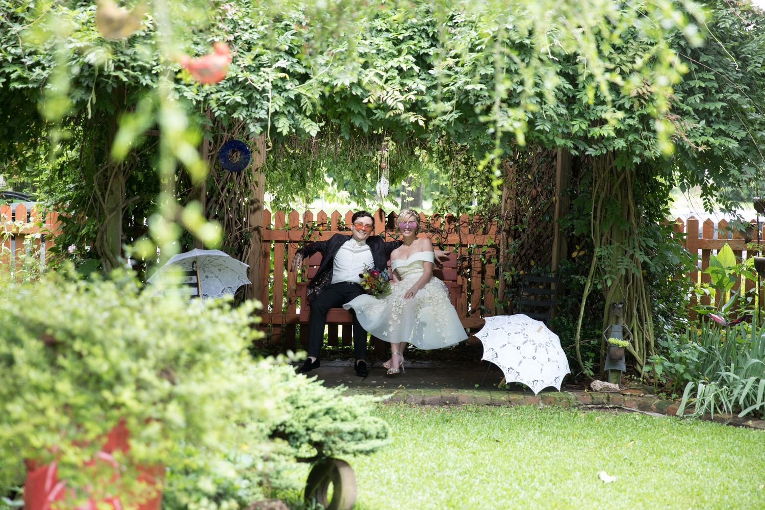 Bride and groom sit together on a garden bench surrounded by lush greenery in a romantic outdoor setting.