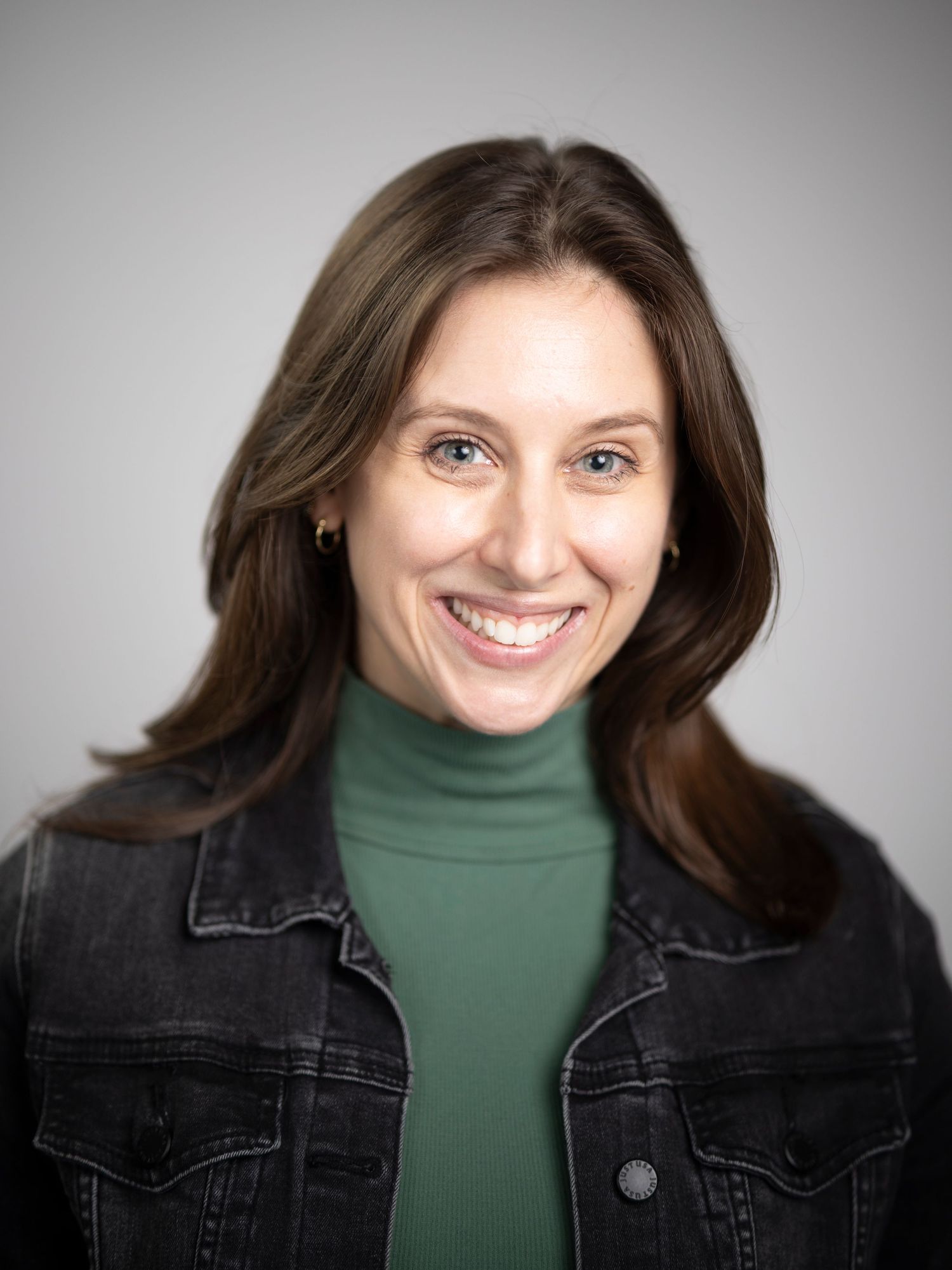 Smiling brunette woman in a green turtleneck and denim jacket posing for a professional headshot.