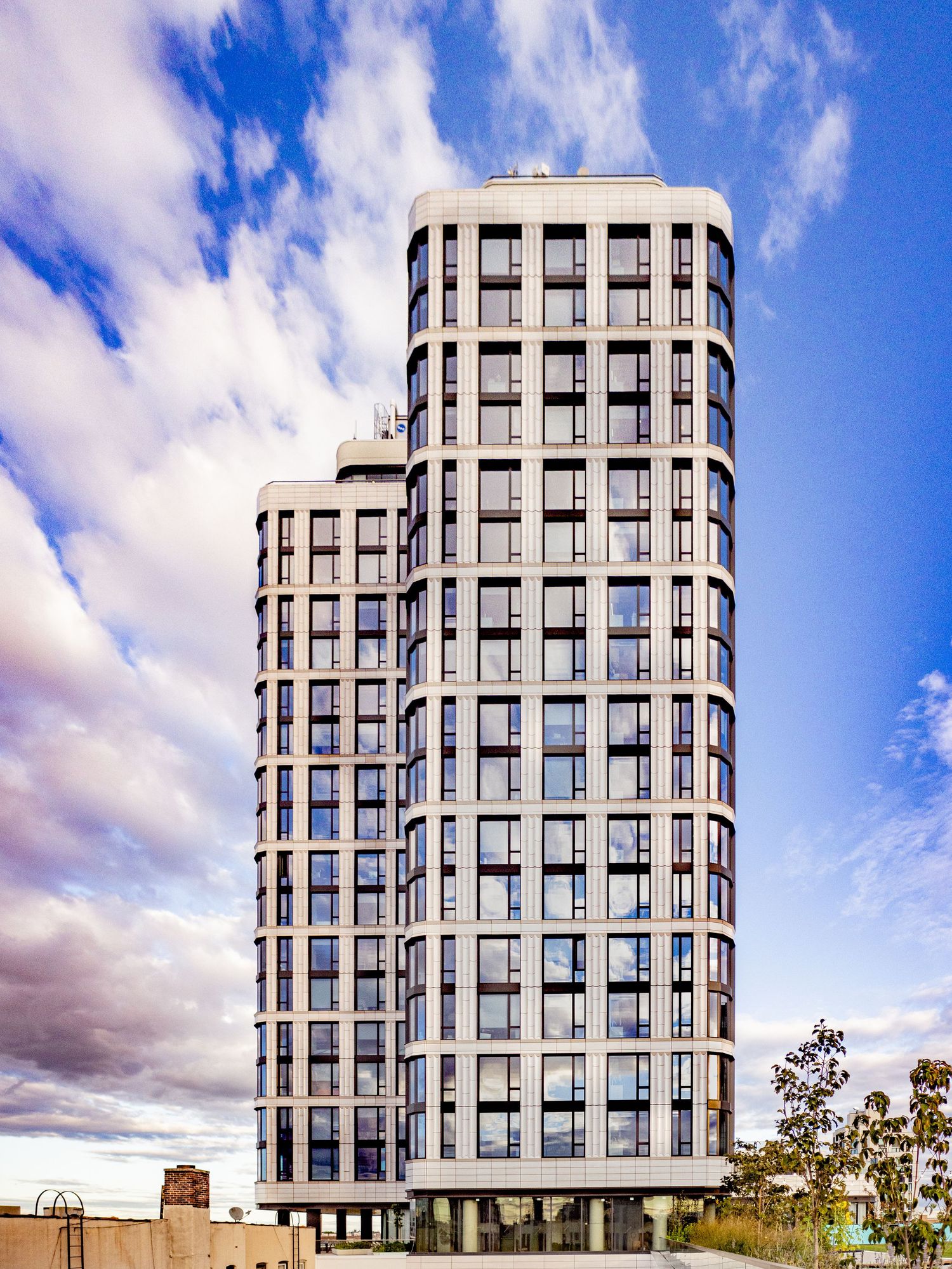 Modern cylindrical residential skyscraper with curved facade and rows of windows against a dramatic blue cloudy sky.