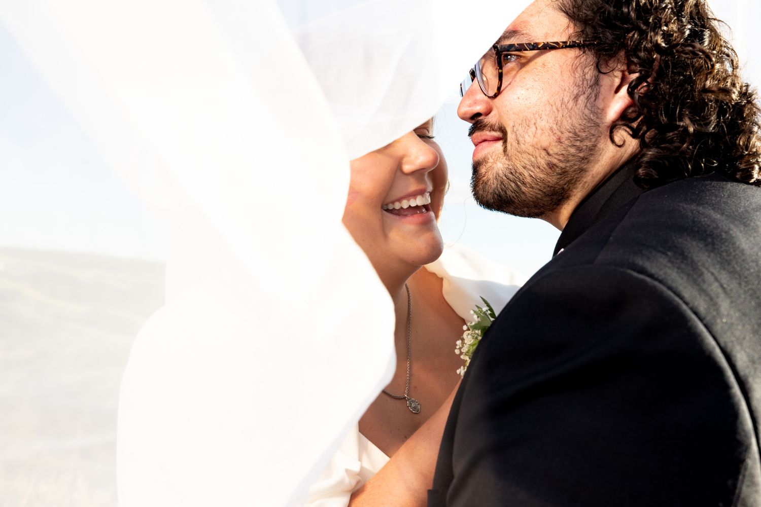 Laughing bride and groom sharing a tender moment under a flowing white veil outdoors.