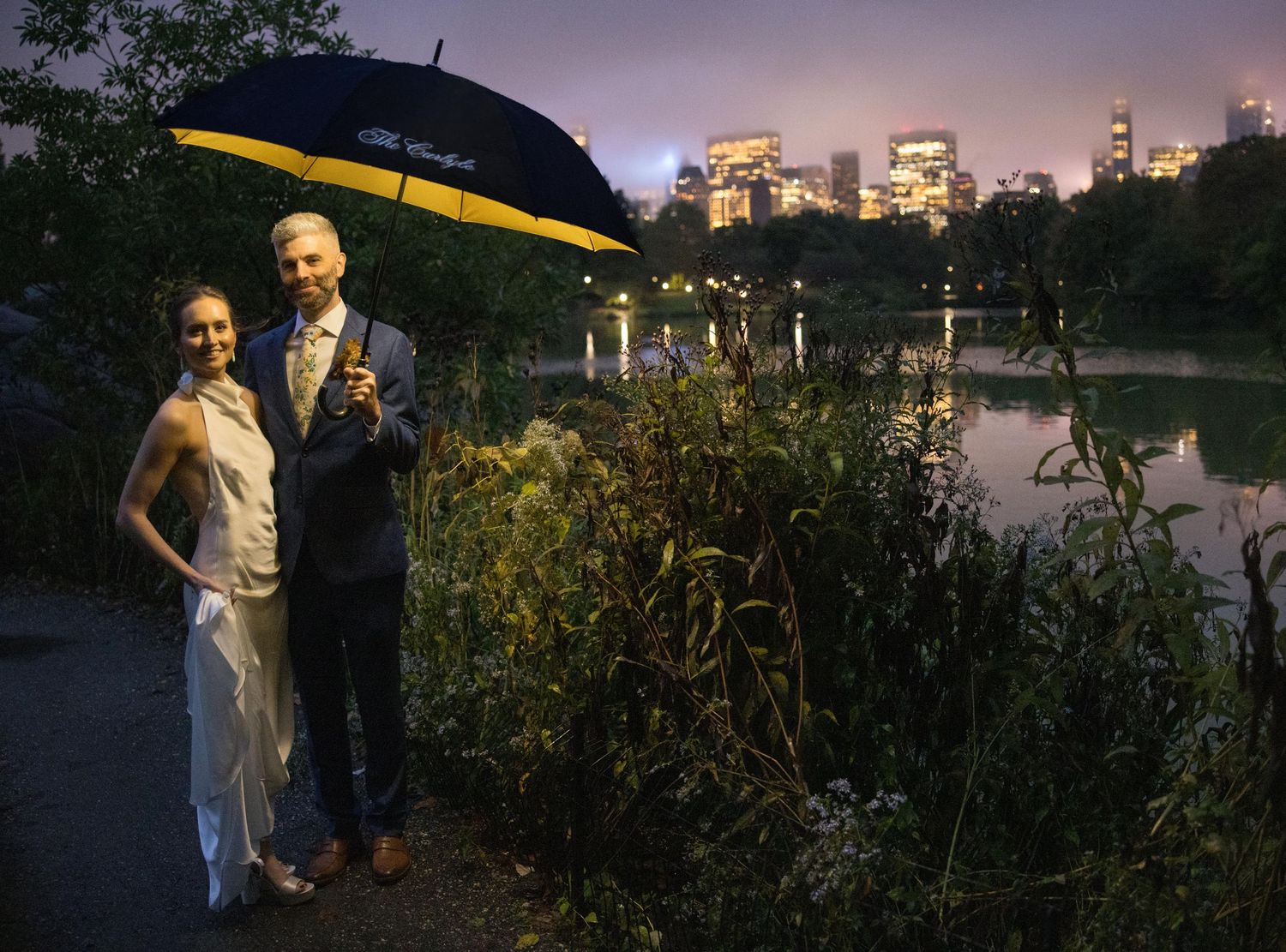 Bride and groom pose under branded umbrella by Central Park lake with glowing NYC skyline at night.