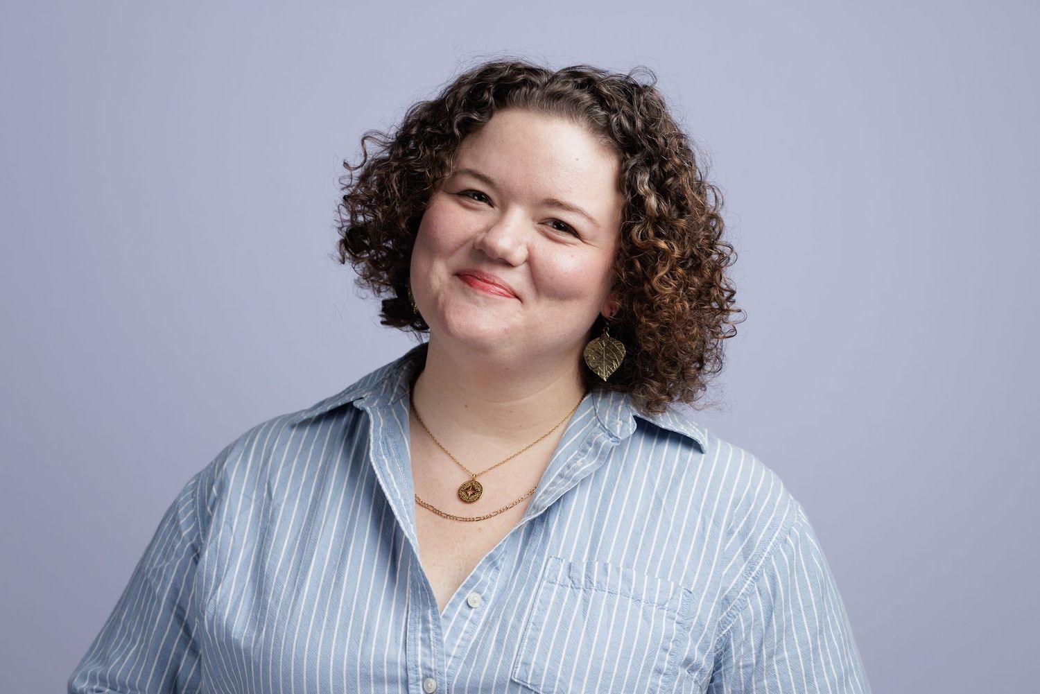 Smiling woman with curly hair wearing a light blue striped shirt and gold necklace posed against a gray background.