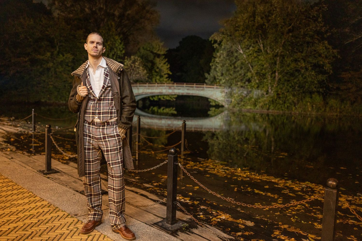 Stylish man in plaid suit standing on pathway near pond at night in autumn park.