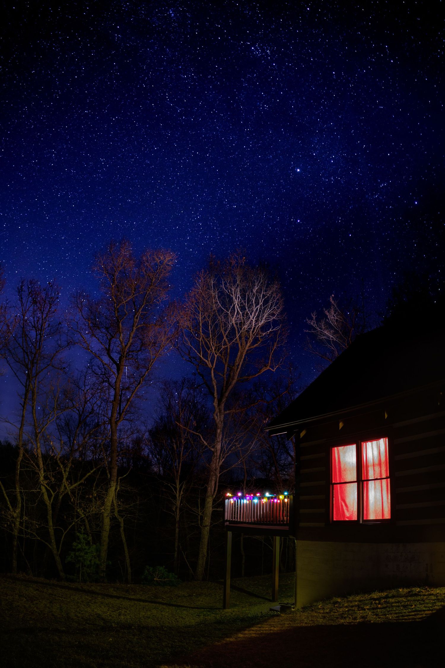 A cozy log cabin with colorful Christmas lights glows warmly under a breathtaking starry night sky surrounded by bare trees.