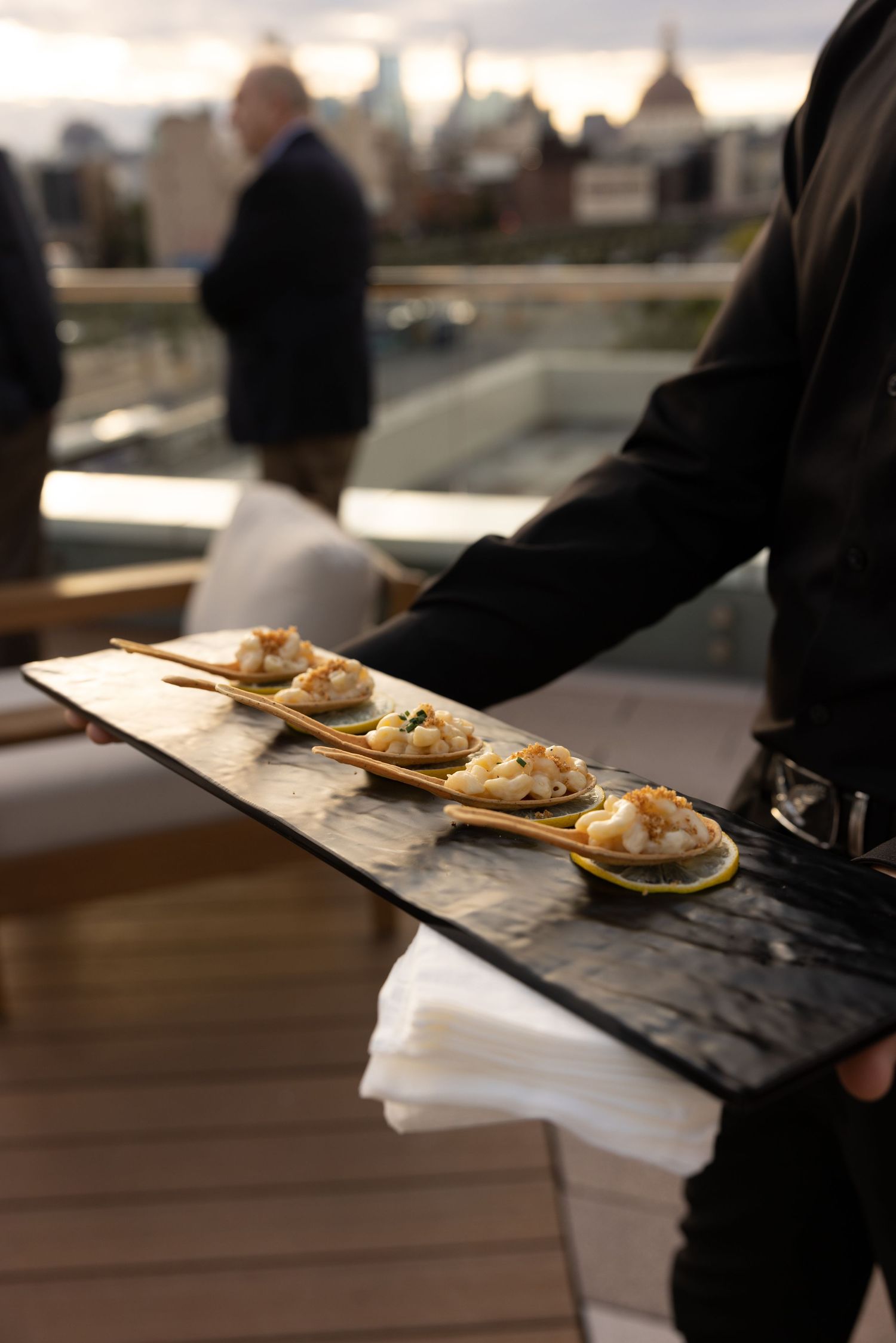 Waiter serving elegant canapés on golden spoons on a slate tray at a rooftop event.