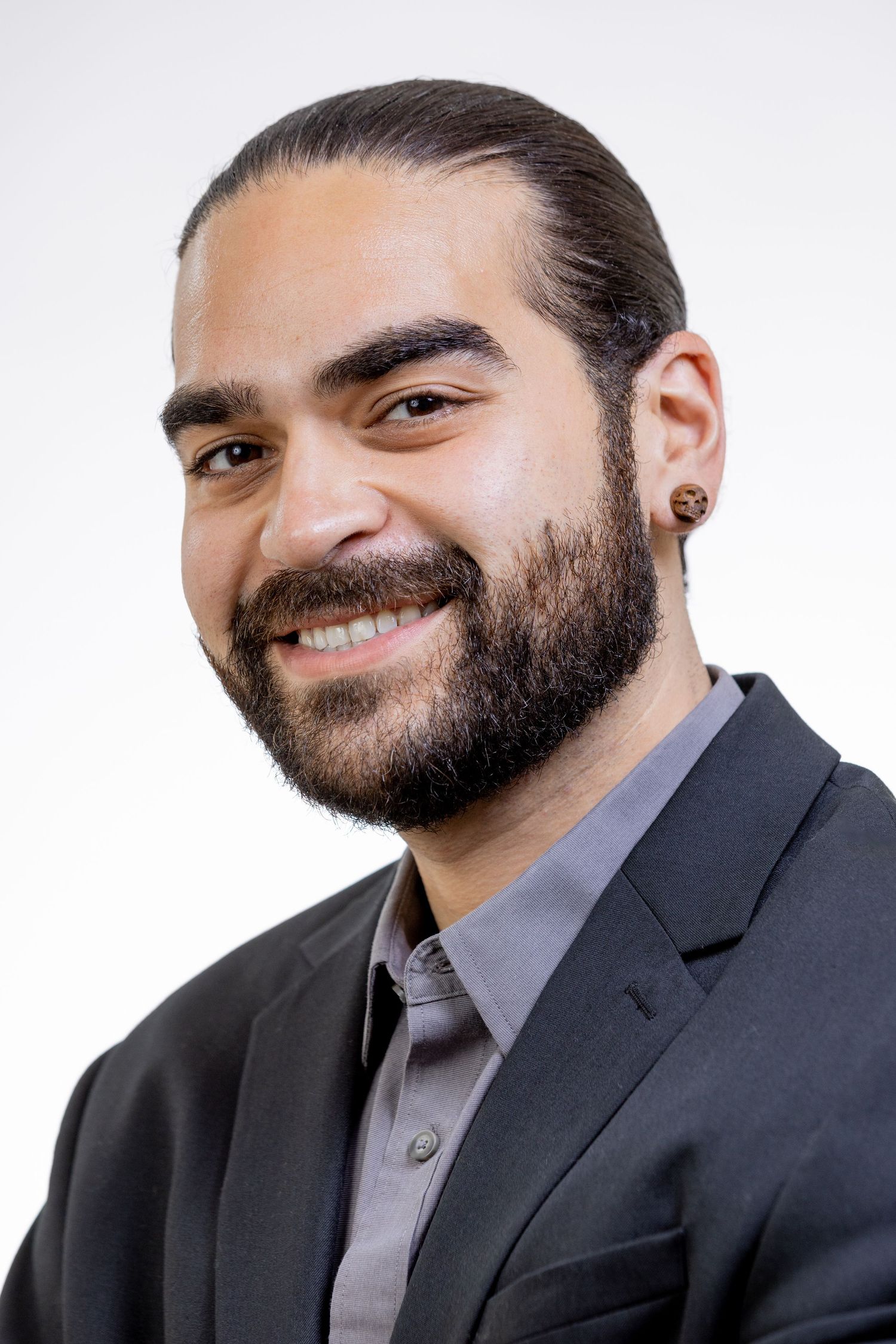 Smiling bearded man in black blazer with ear gauges against white background.