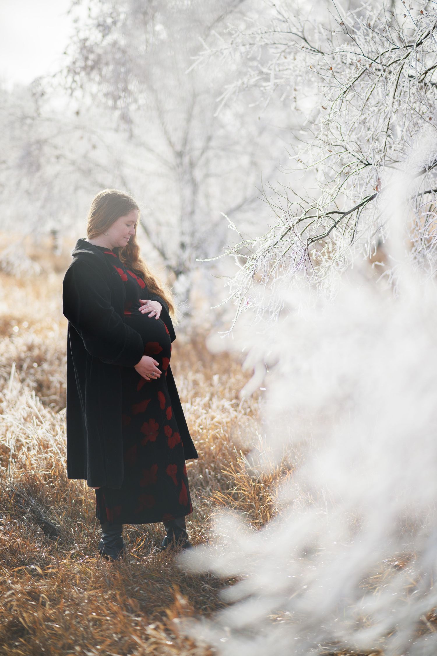 Pregnant woman in black coat cradling belly outdoors in a frost-covered winter landscape.