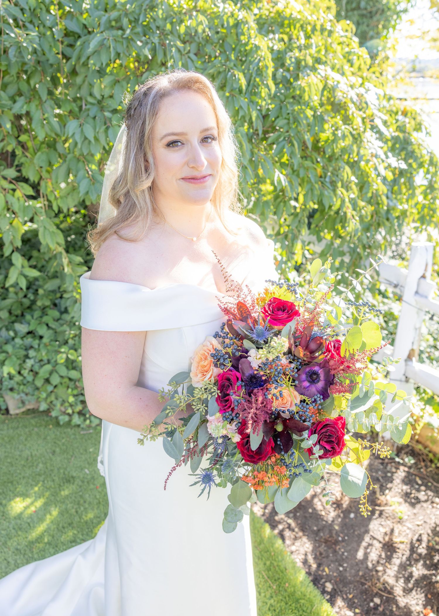 Bride in off-shoulder white gown holds vibrant bouquet of red, purple, and orange flowers outdoors.