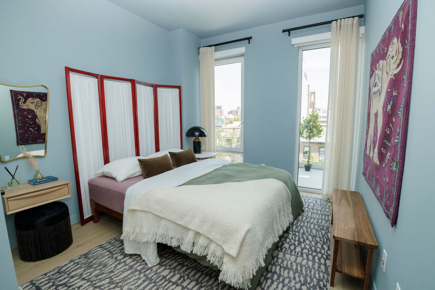 Modern bedroom with blue walls, red-framed wardrobe, large bed, and balcony access with city views.