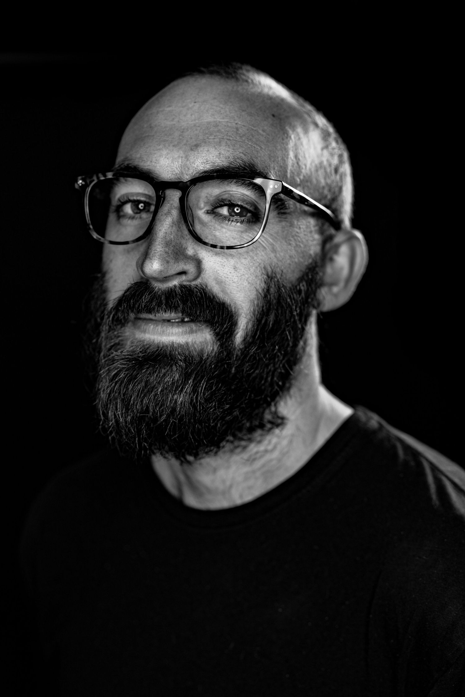 Black and white portrait of bald bearded white man wearing glasses on dark background.