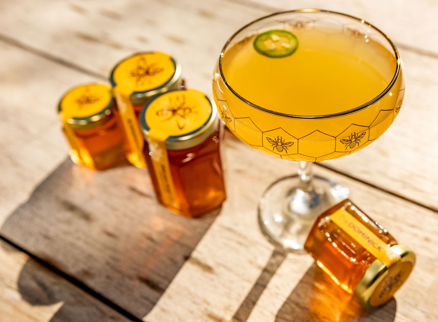 Golden honey cocktail in a coupe glass garnished with jalapeno, served alongside honey jar favors.
