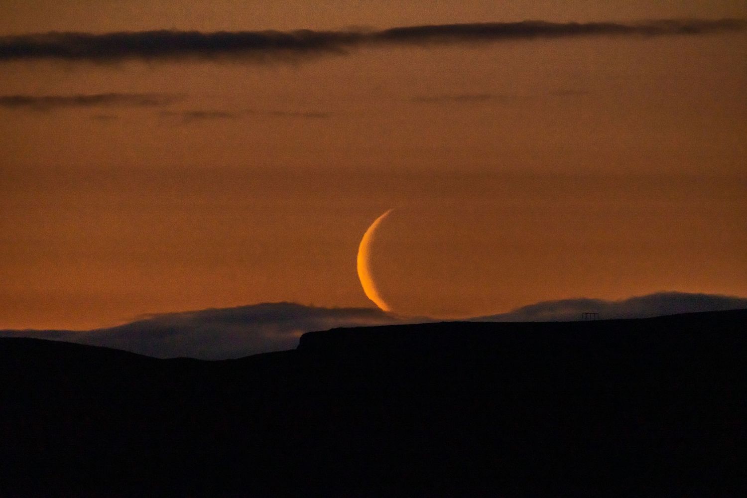 Crescent moon glowing orange against a deep amber sunset sky above dark silhouetted mountain ridges.