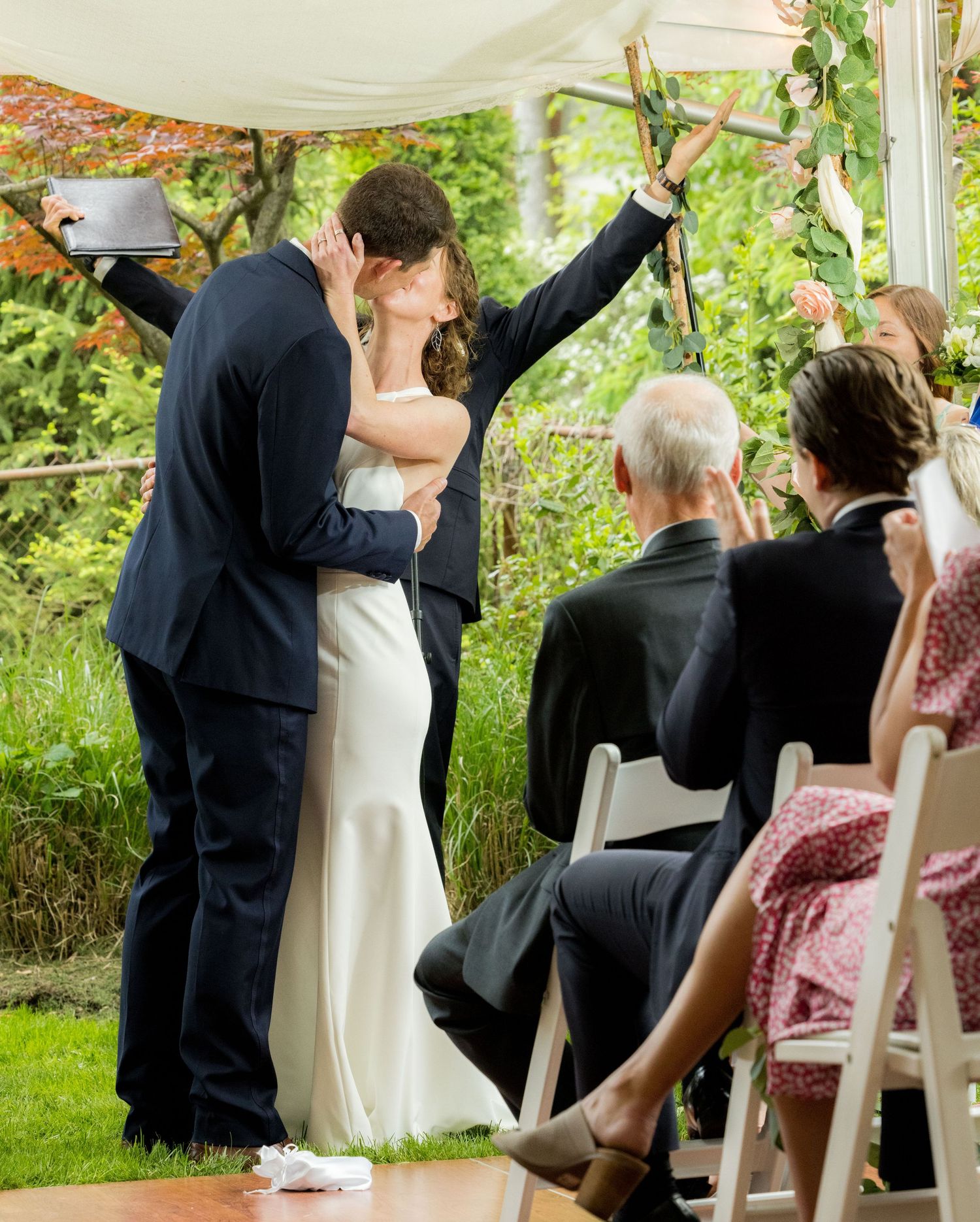 Bride and groom share first kiss at outdoor garden wedding ceremony under floral canopy.