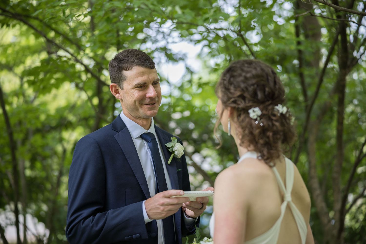 Groom in navy suit smiling while bride reads wedding vows outdoors surrounded by lush green trees.