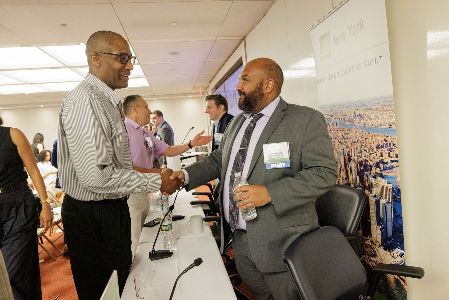 Two men shake hands and converse at a professional networking event near a display table.