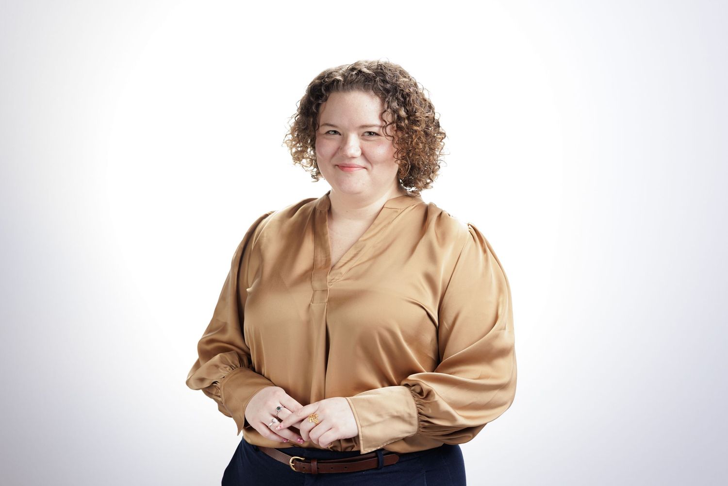 Professional woman with curly hair wearing a gold satin blouse, smiling confidently against a white background.