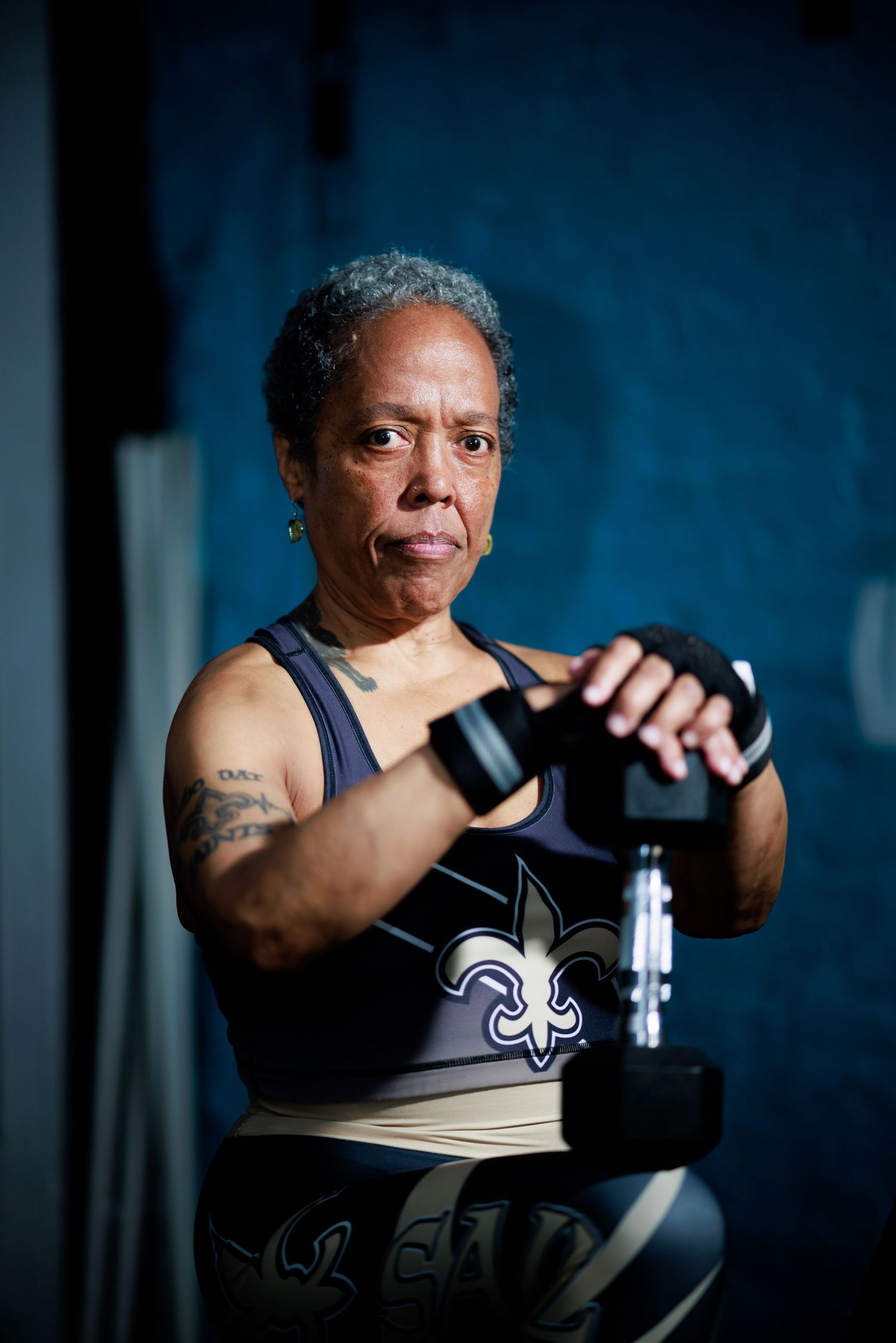 Strong older Black woman in Saints sports bra focused on lifting dumbbells during an intense gym workout.