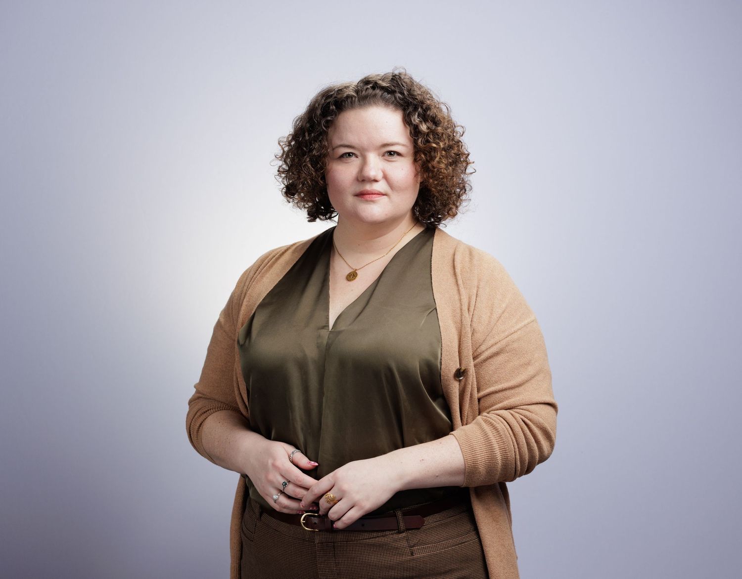 Professional woman with curly hair wearing olive blouse and tan cardigan posing confidently against gray background.