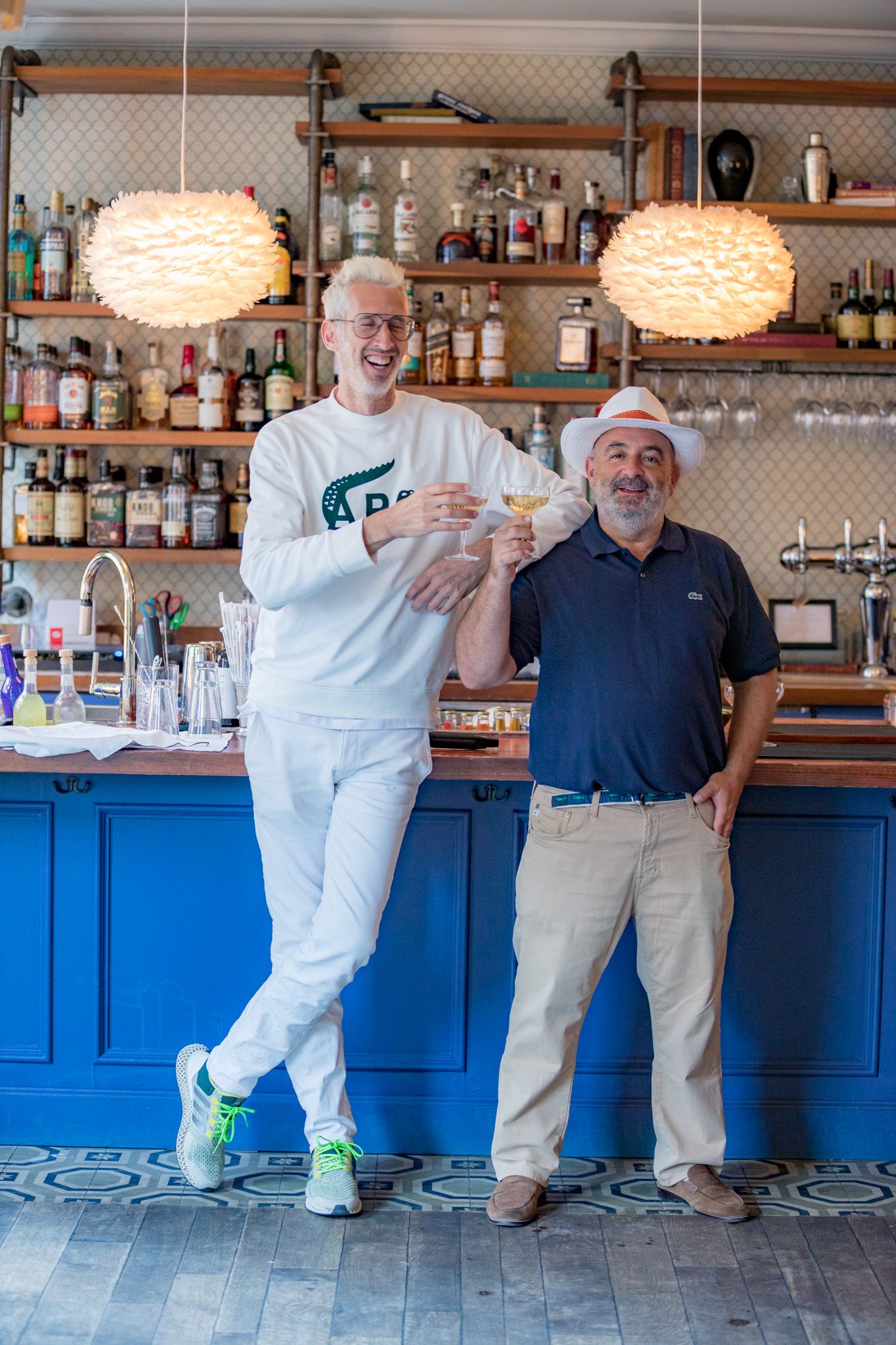 Two men smiling and toasting drinks at a blue bar with shelves of liquor bottles behind them.