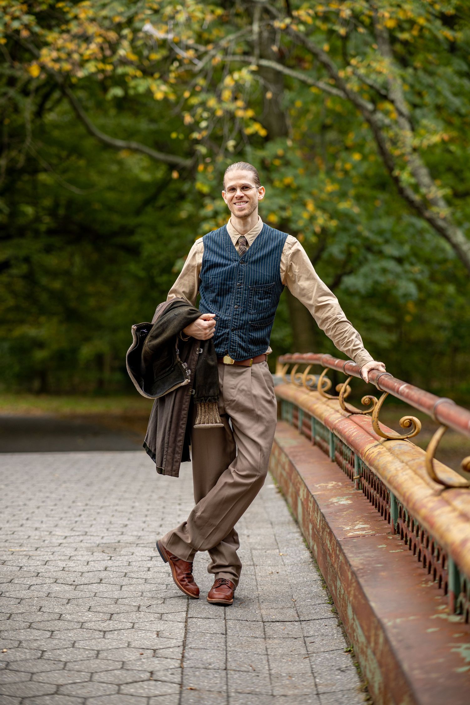 Smiling man in vest and tie holding jacket on a decorative bridge in an autumn park.