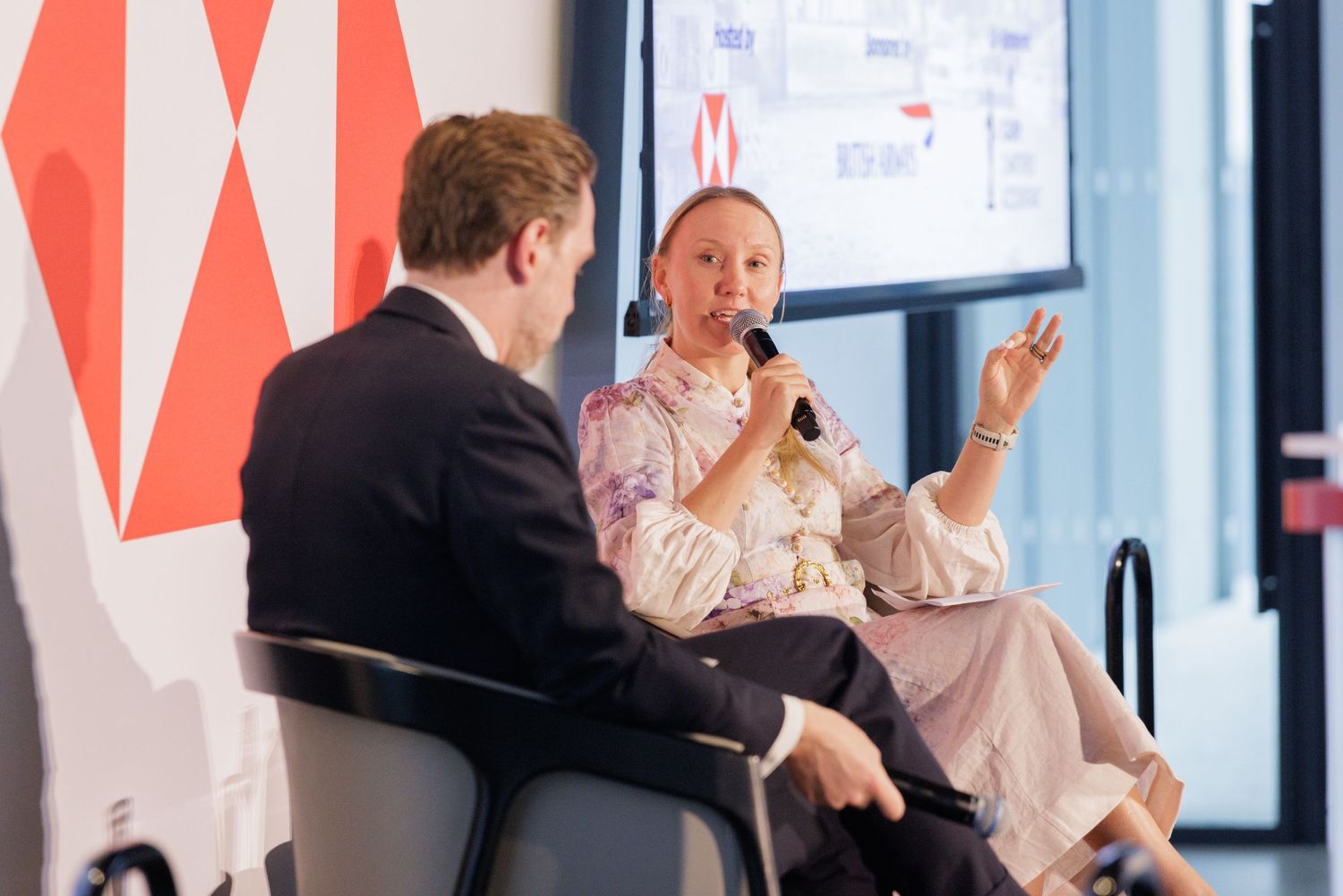 Woman speaking into microphone during a panel discussion at a modern corporate event with red geometric decor.