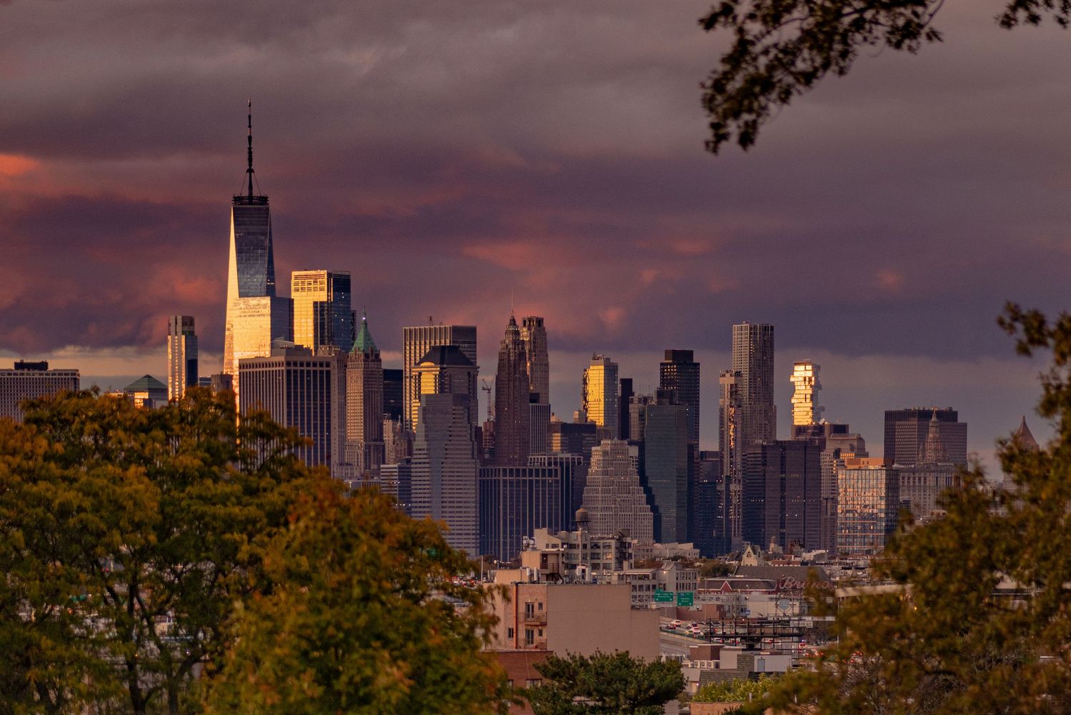 Dramatic sunset view of Lower Manhattan skyline with One World Trade Center framed by autumn trees from Brooklyn.