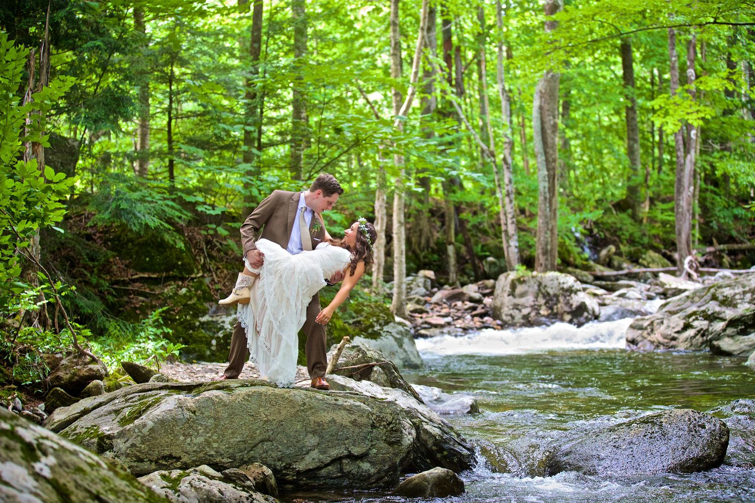 Groom carrying bride in white wedding dress over rocks beside a rushing forest stream.
