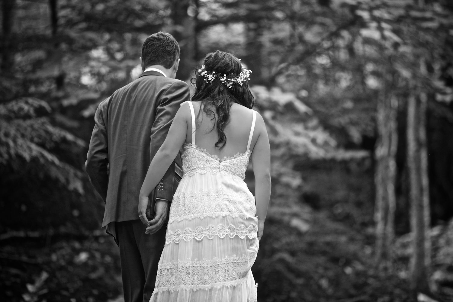Black and white photo of bride in lace gown with flower crown walking with groom through forest.