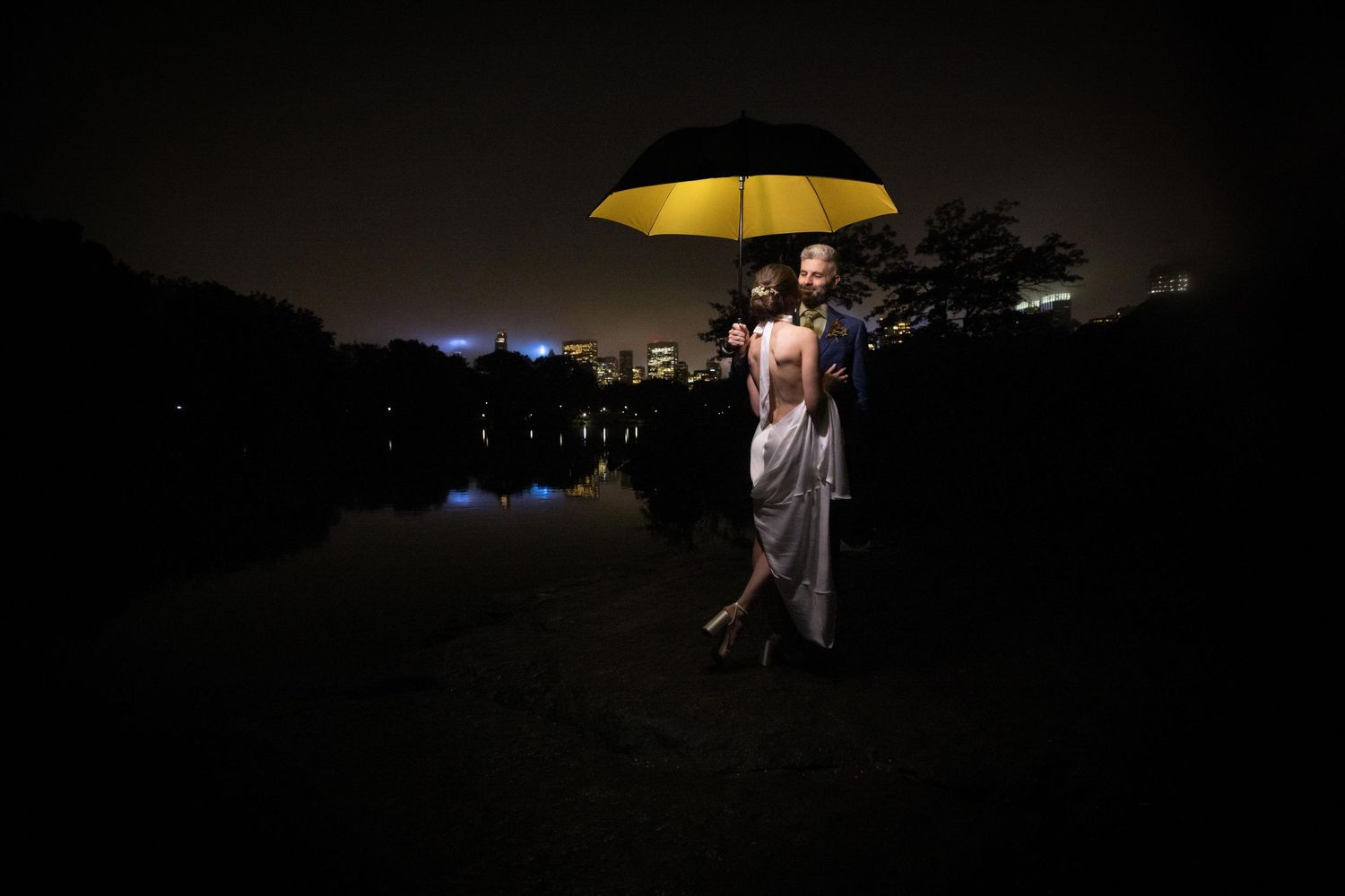 Bride and groom stand romantically under a glowing yellow umbrella beside a reflective lake at night.