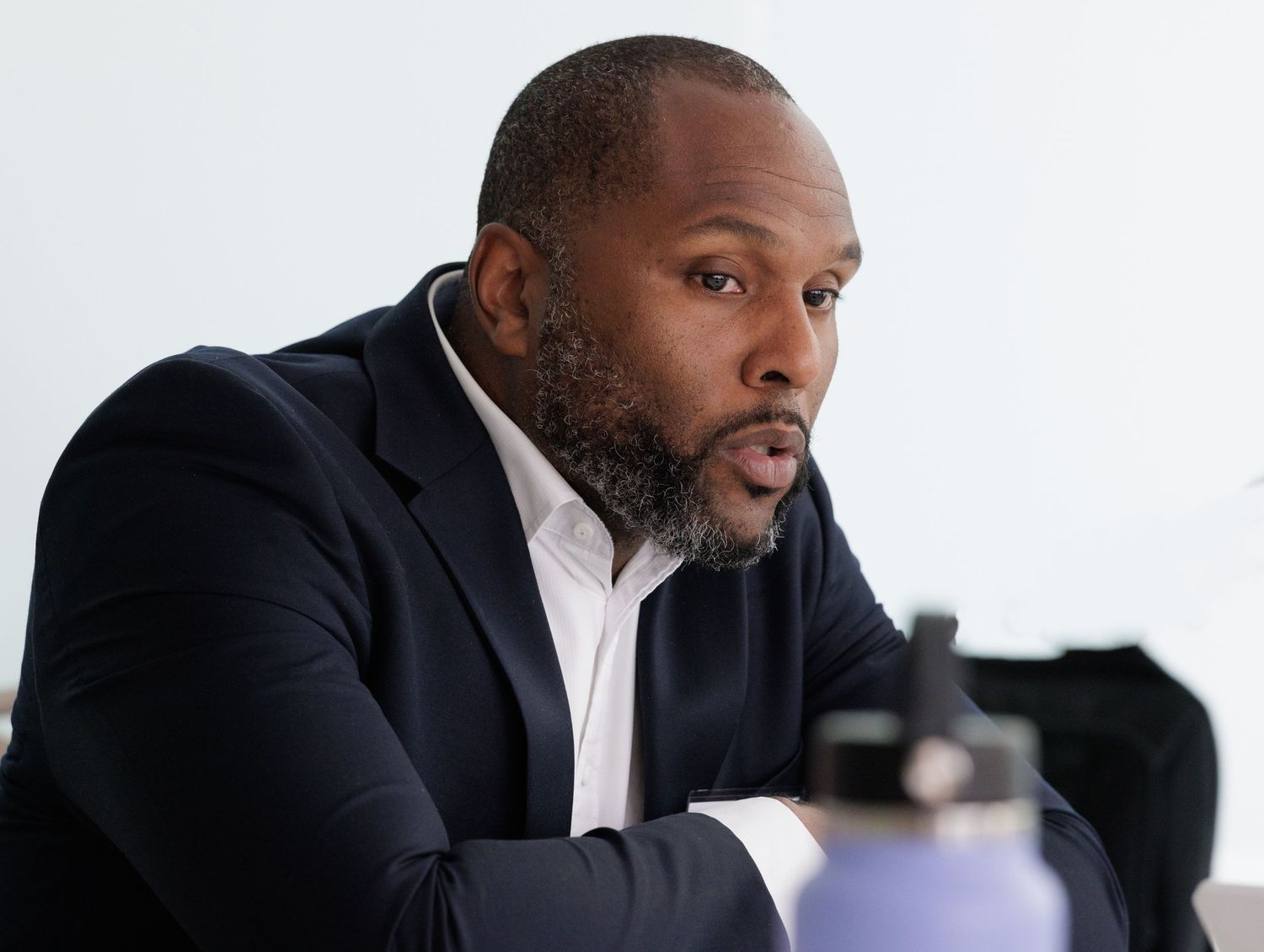 A Black man in a dark blazer sits leaning forward during a professional meeting or conference.