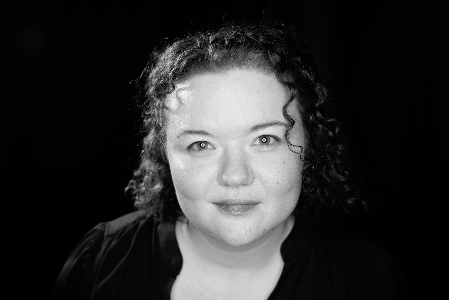 Black and white portrait of a curly-haired woman in a dark top, smiling softly against a dark background.