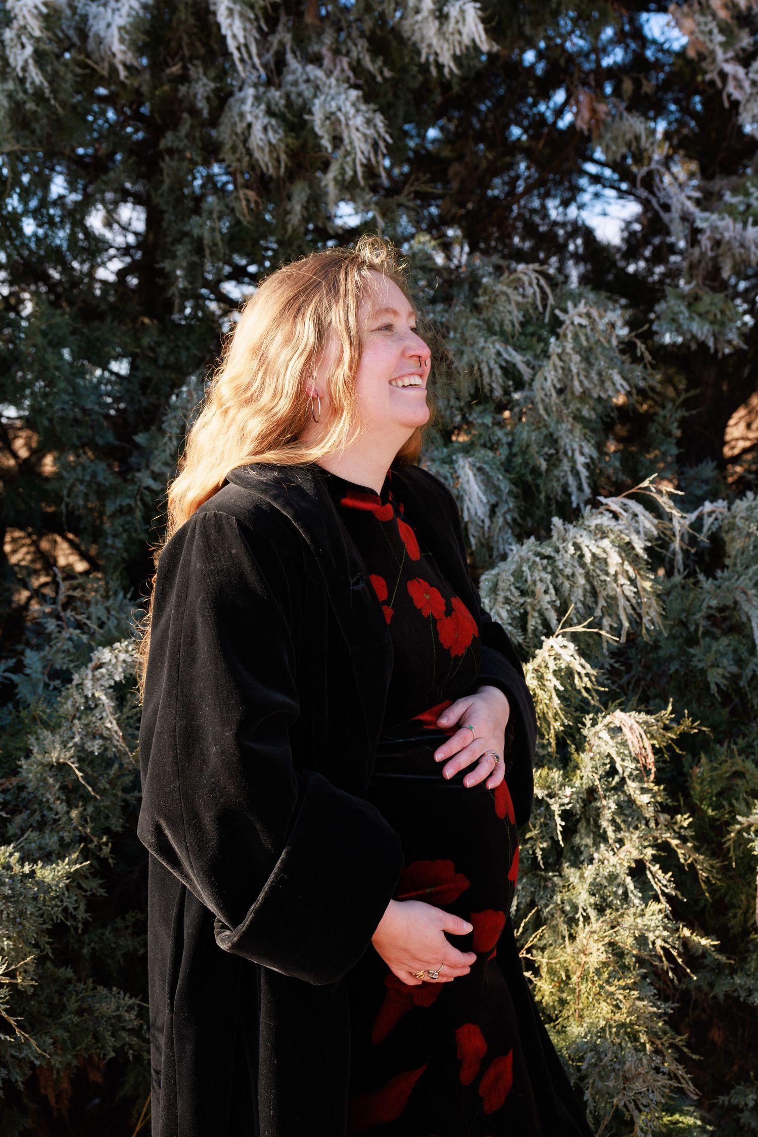 Smiling pregnant woman in black velvet coat and red floral dress cradling her baby bump outdoors near evergreen trees.