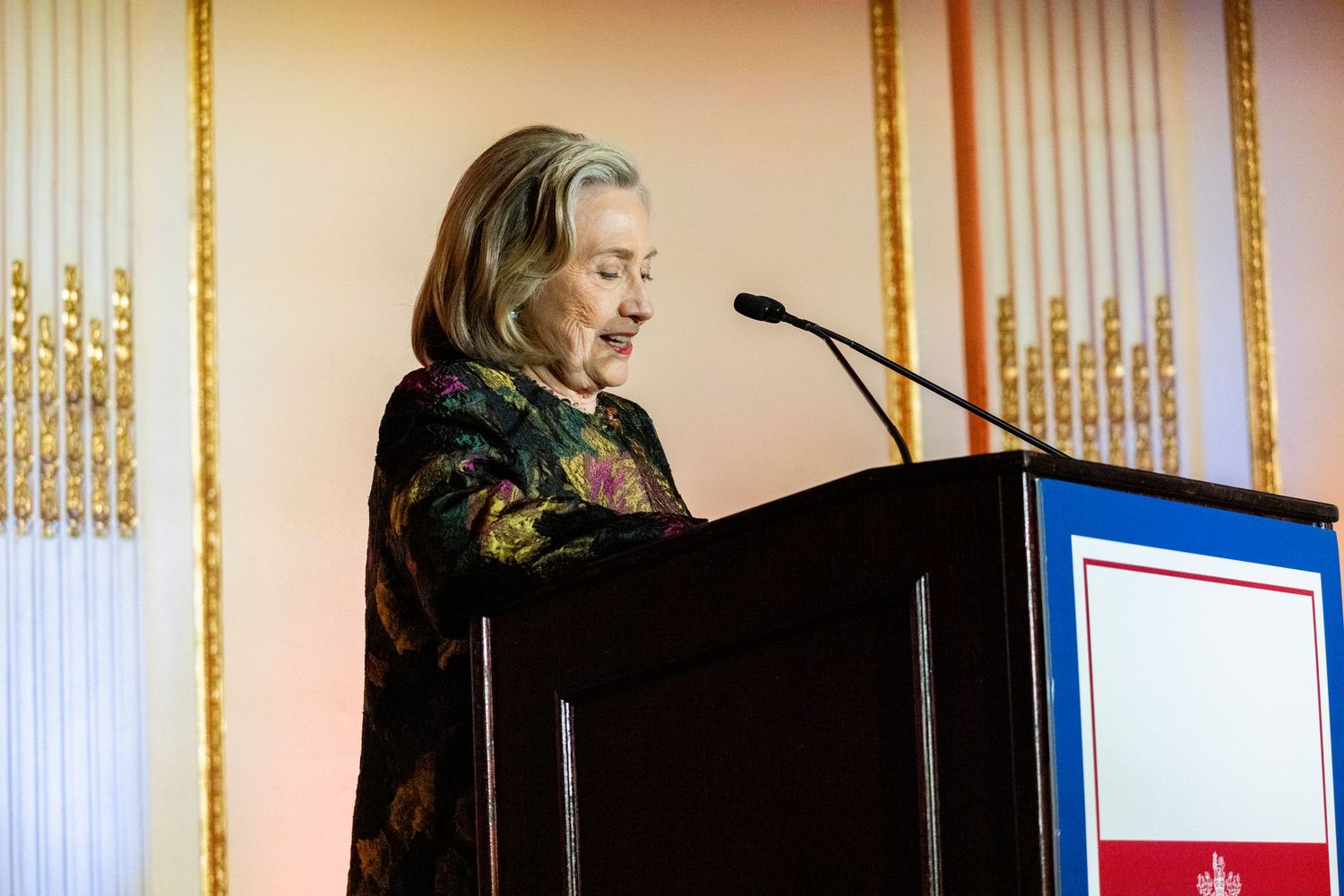 Woman in floral dress speaking at a podium during a formal awards ceremony or gala event.