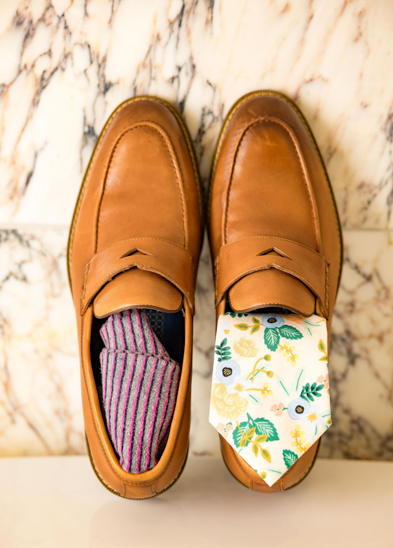 Brown leather penny loafers displayed on marble surface with floral tie and striped socks for groom's attire.