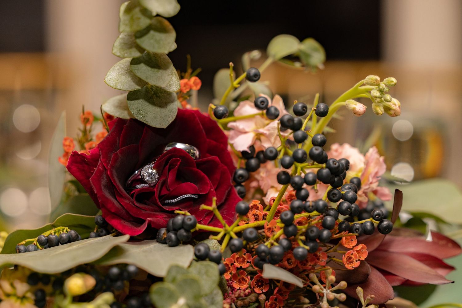 Elegant dark floral wedding bouquet with deep red roses, berries, and greenery with rings placed on top.