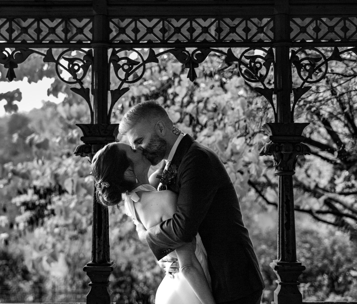 Bride and groom sharing a romantic kiss under an ornate gazebo in black and white.